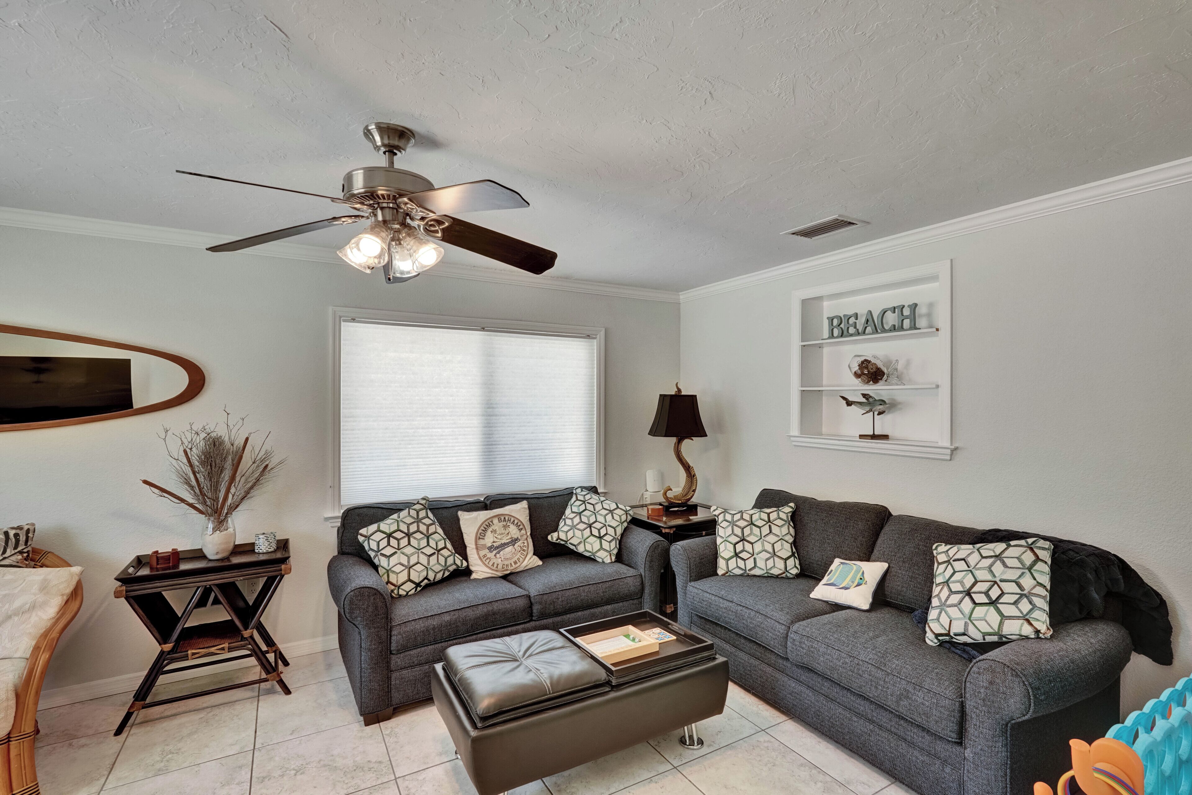 A living room with a ceiling fan, a sofa set, a coffee table, a side table with a lamp, and a wall-mounted shelf with decorative items.
