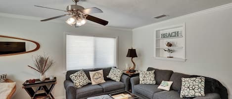 A living room with a ceiling fan, a sofa set, a coffee table, a side table with a lamp, and a wall-mounted shelf with decorative items.