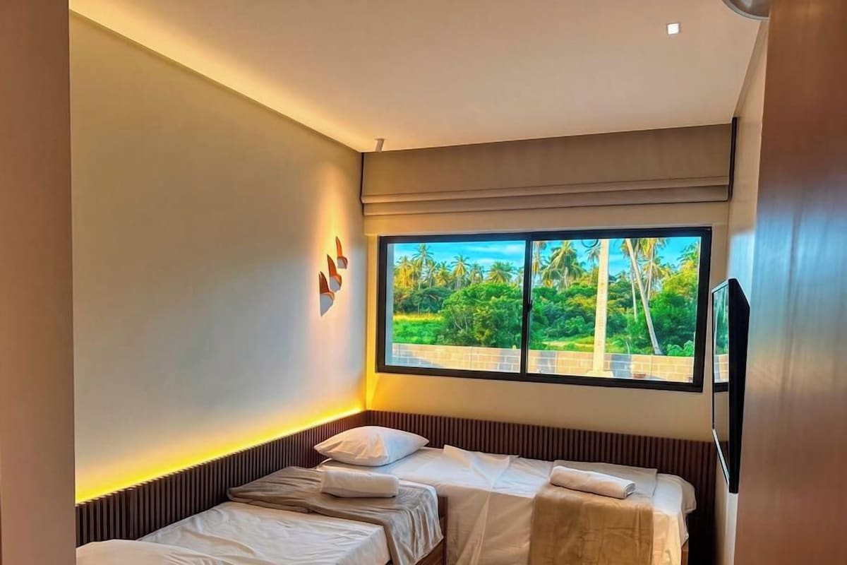 A hotel room with two beds, a large window with a view of greenery, and a flat-screen TV mounted on the wall.