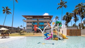 A swimming pool with slides and a play structure, surrounded by palm trees and a building with multiple balconies. - Beachfront Condos at Ipioca Beach Life Maceio by MME (Maceió)