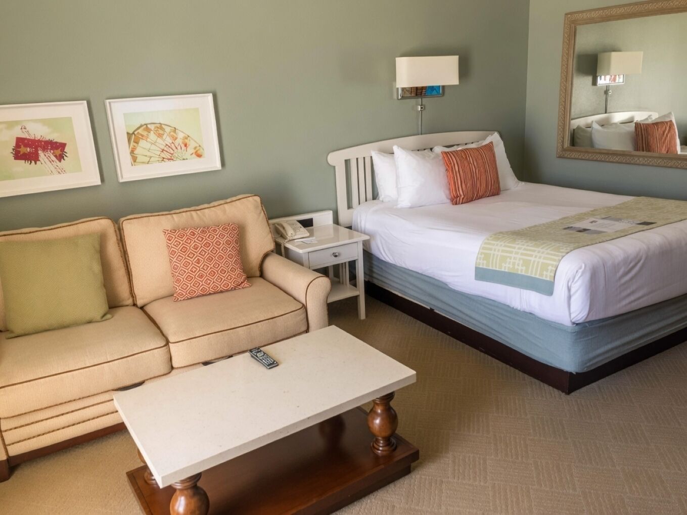 Habitación de hotel con cama, sofá, mesita de noche, lámpara y dos cuadros en la pared.