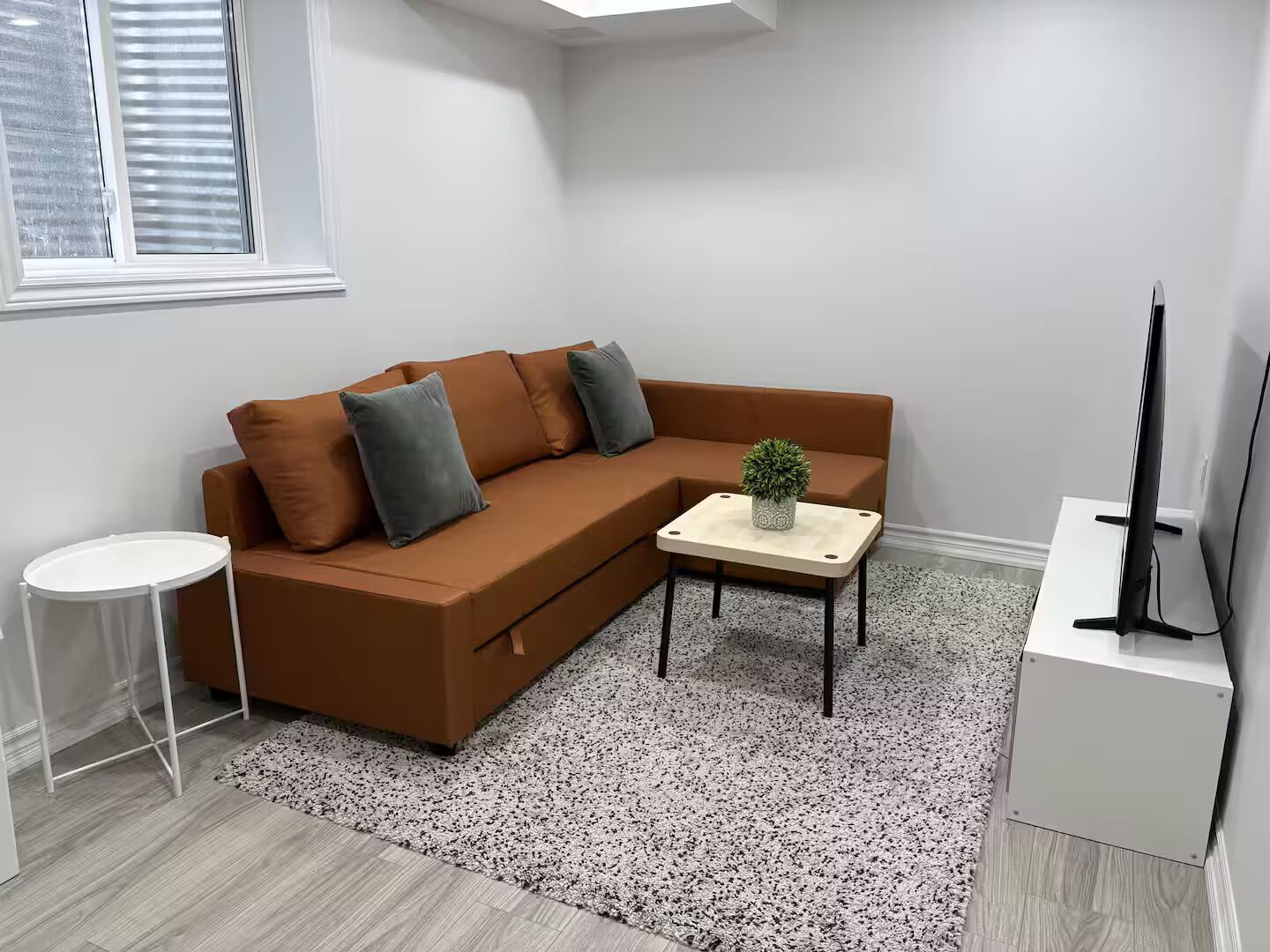A modern living room with a brown sectional sofa, a small white side table, a coffee table with a plant, a TV stand, and a window with blinds.