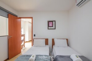 Quarto com duas camas, porta de madeira, pequena moldura na parede e uma toalha sobre uma das camas.
