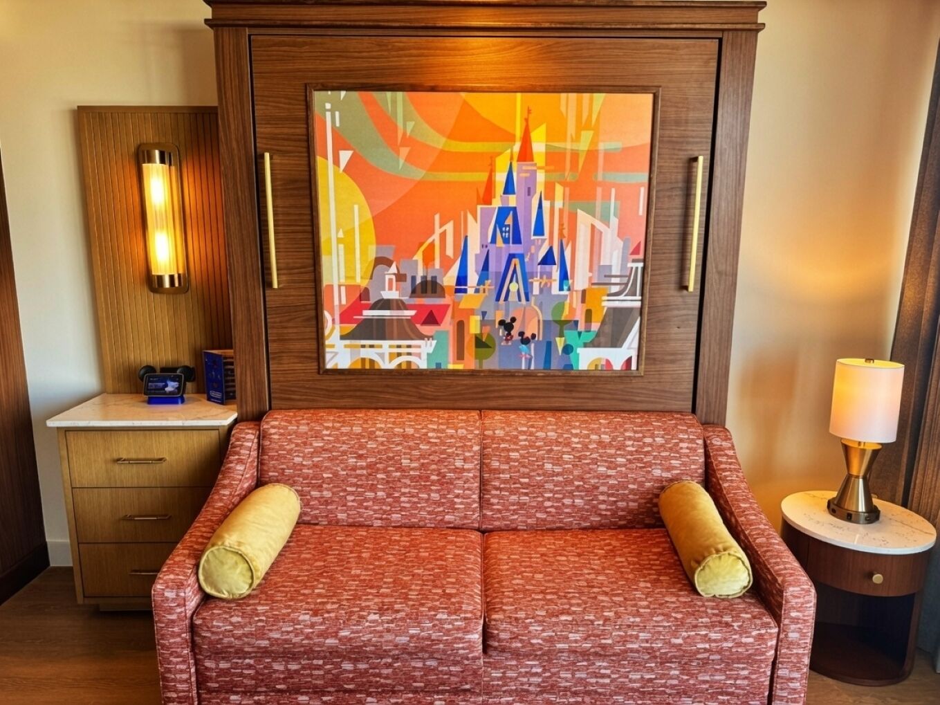 A hotel room with a red patterned sofa, a wooden cabinet, a wall-mounted painting, a bedside lamp, and a small bedside table.