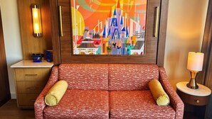 Quarto de hotel com um sofá estampado em vermelho, um armário de madeira, um quadro pendurado na parede, um abajur e uma mesinha de cabeceira.
