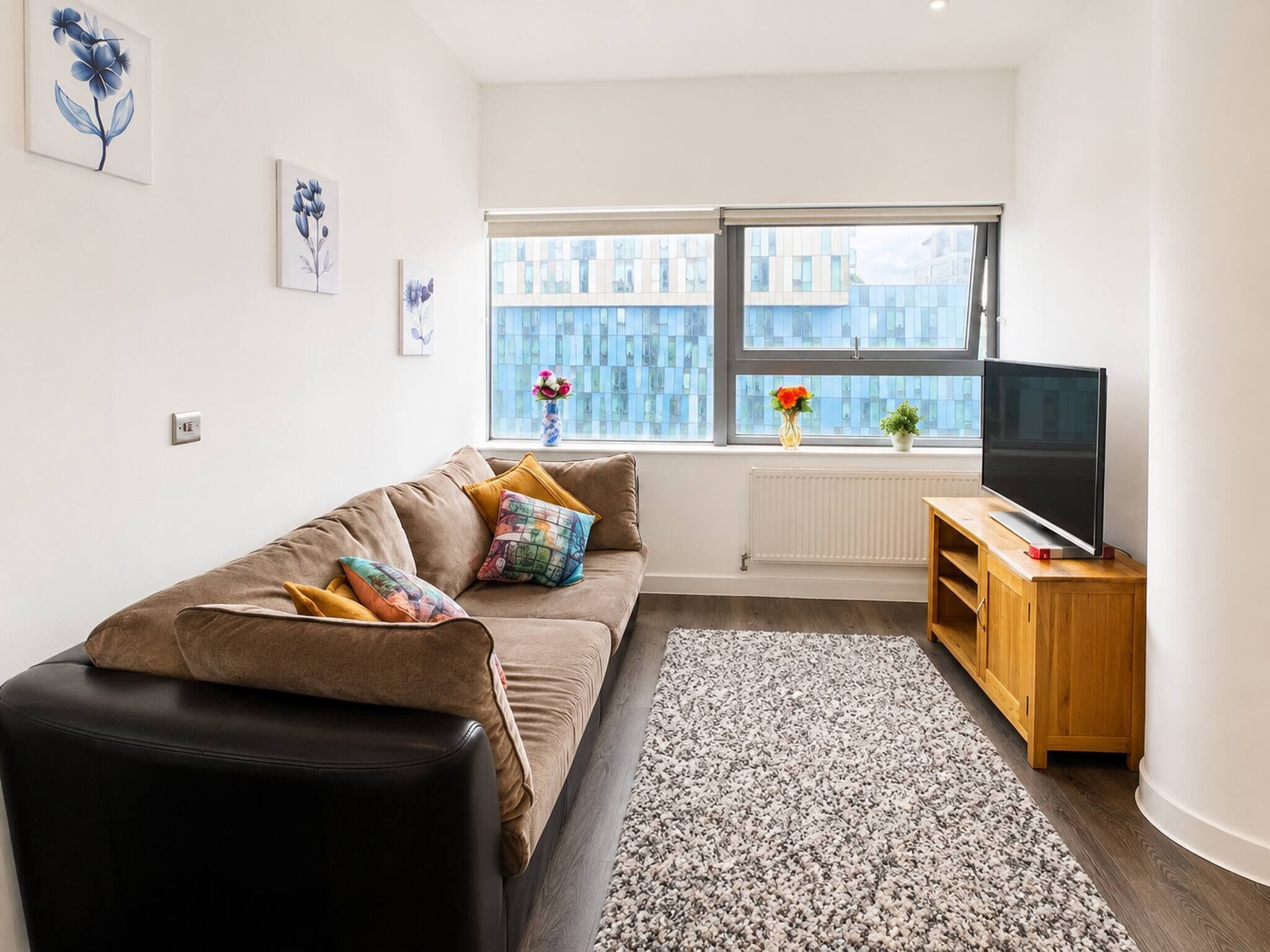 Ein modernes Wohnzimmer mit einer Couch, einem Flachbildfernseher, einem Holzschrank und einem Fenster mit Blick auf Gebäude.