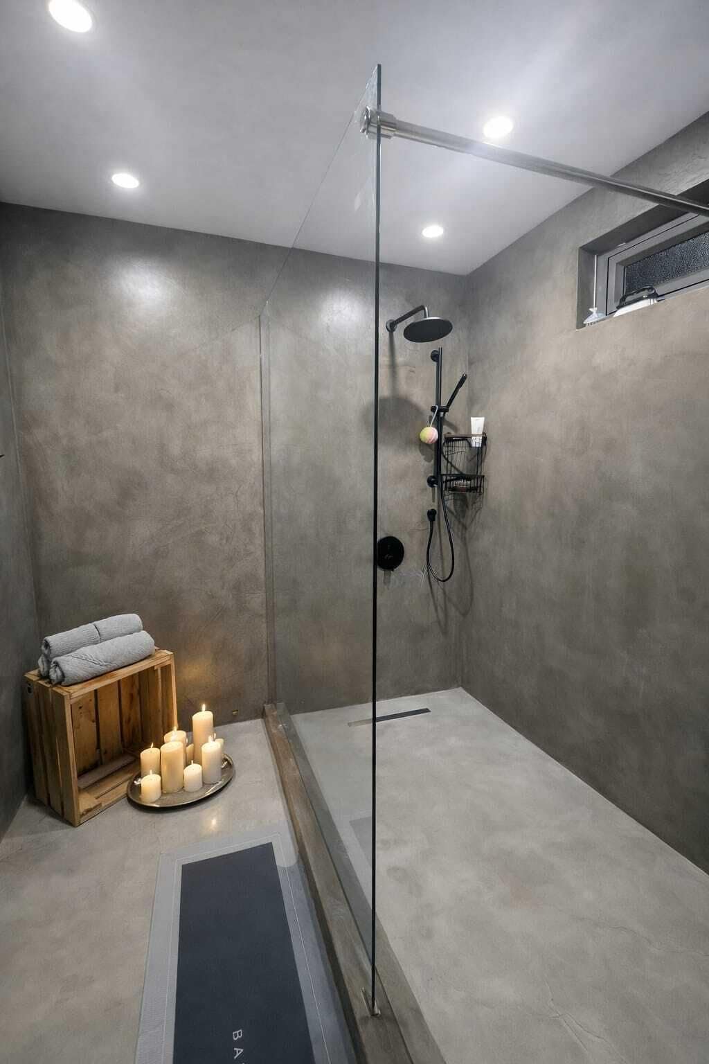 A modern shower area with a glass door, a wooden bench with towels, and candles on the floor.