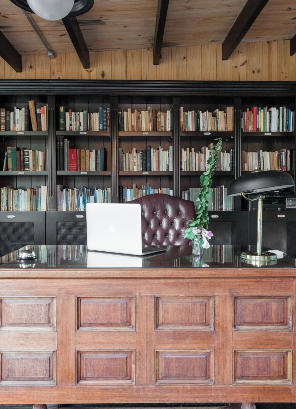 A wooden desk with a laptop, a leather chair, a vase with flowers, and a bookshelf filled with books.