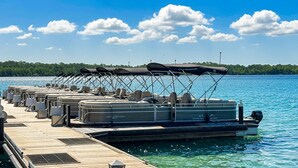 Eine Anlegestelle mit mehreren Pontonbooten, ausgestattet mit Sonnensegeln, einem Motorboot und freiem Blick auf das Wasser und die Küste in der Ferne.
