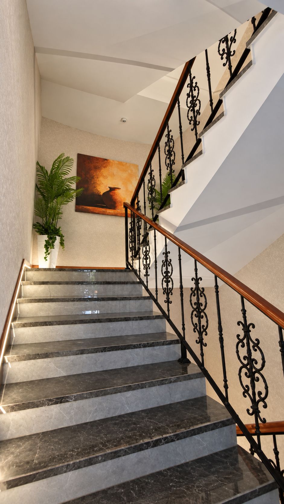 A marble staircase with ornate black railings and a wooden handrail, leading to a landing with a framed artwork and a potted plant.