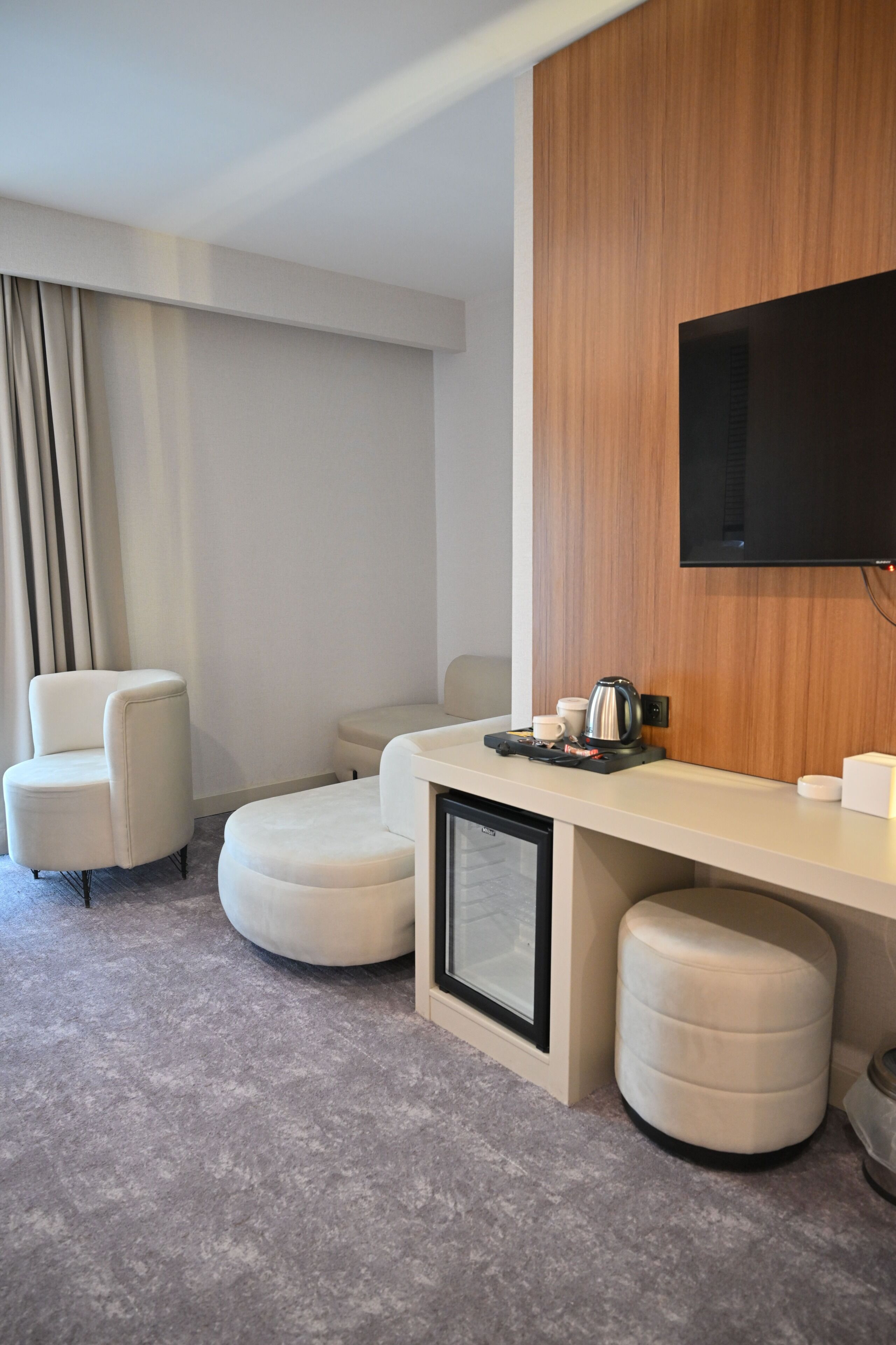 Habitación de hotel moderna con televisor de pantalla plana montado en un panel de madera, un sofá, una mesita con un hervidor y una máquina de café.