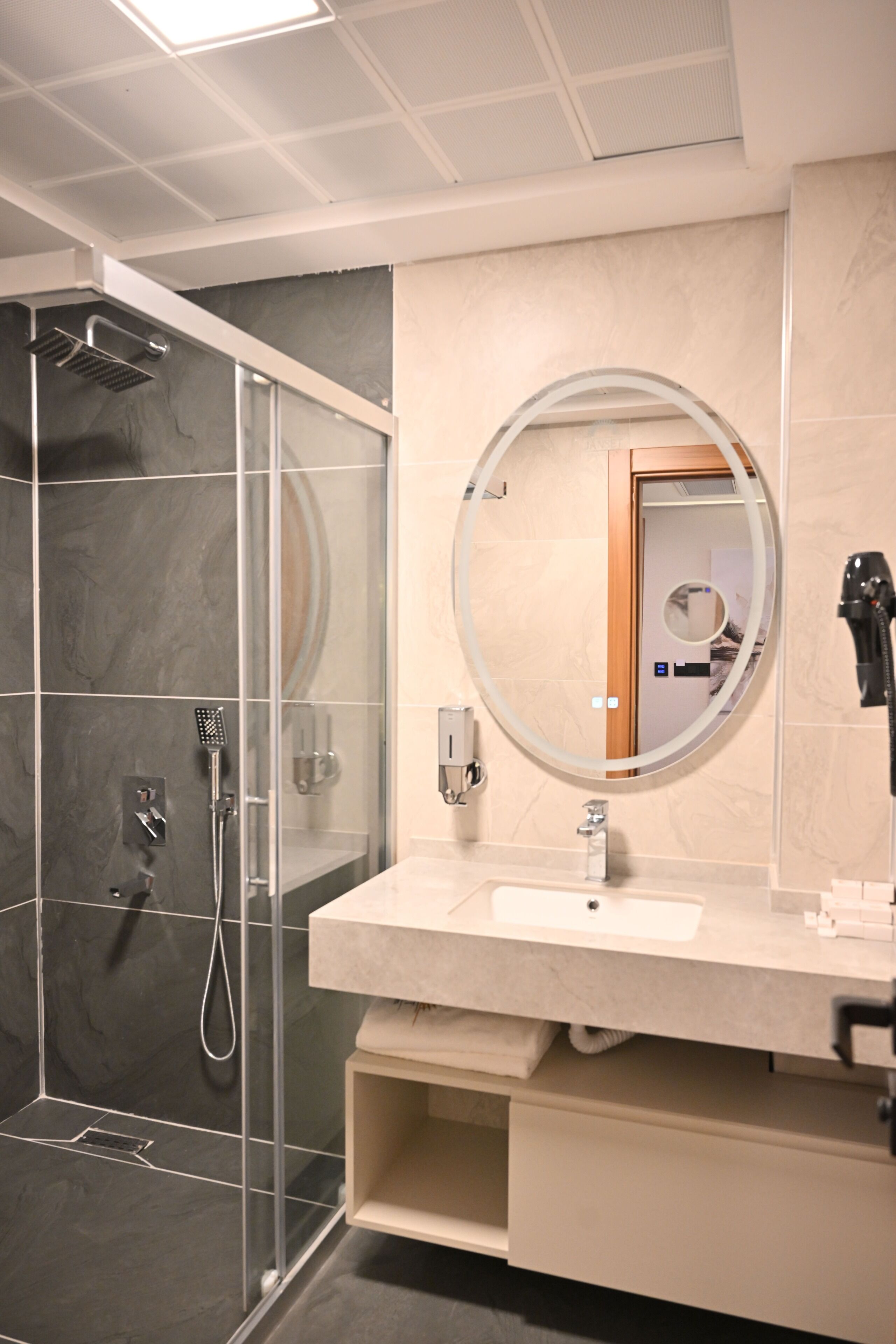 A modern bathroom with a glass-enclosed shower, a round mirror, and a beige vanity with a sink.