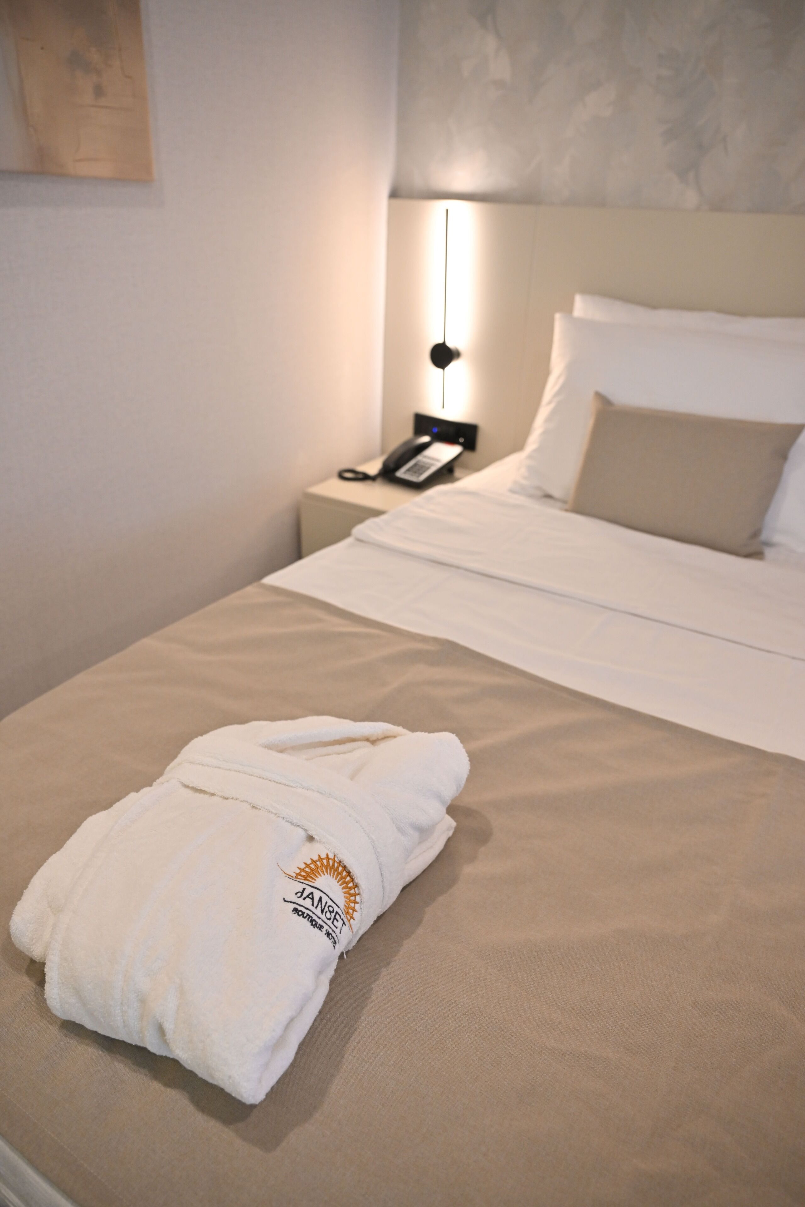 A neatly made bed with a white robe on it, a bedside table with a phone, and a wall-mounted lamp.