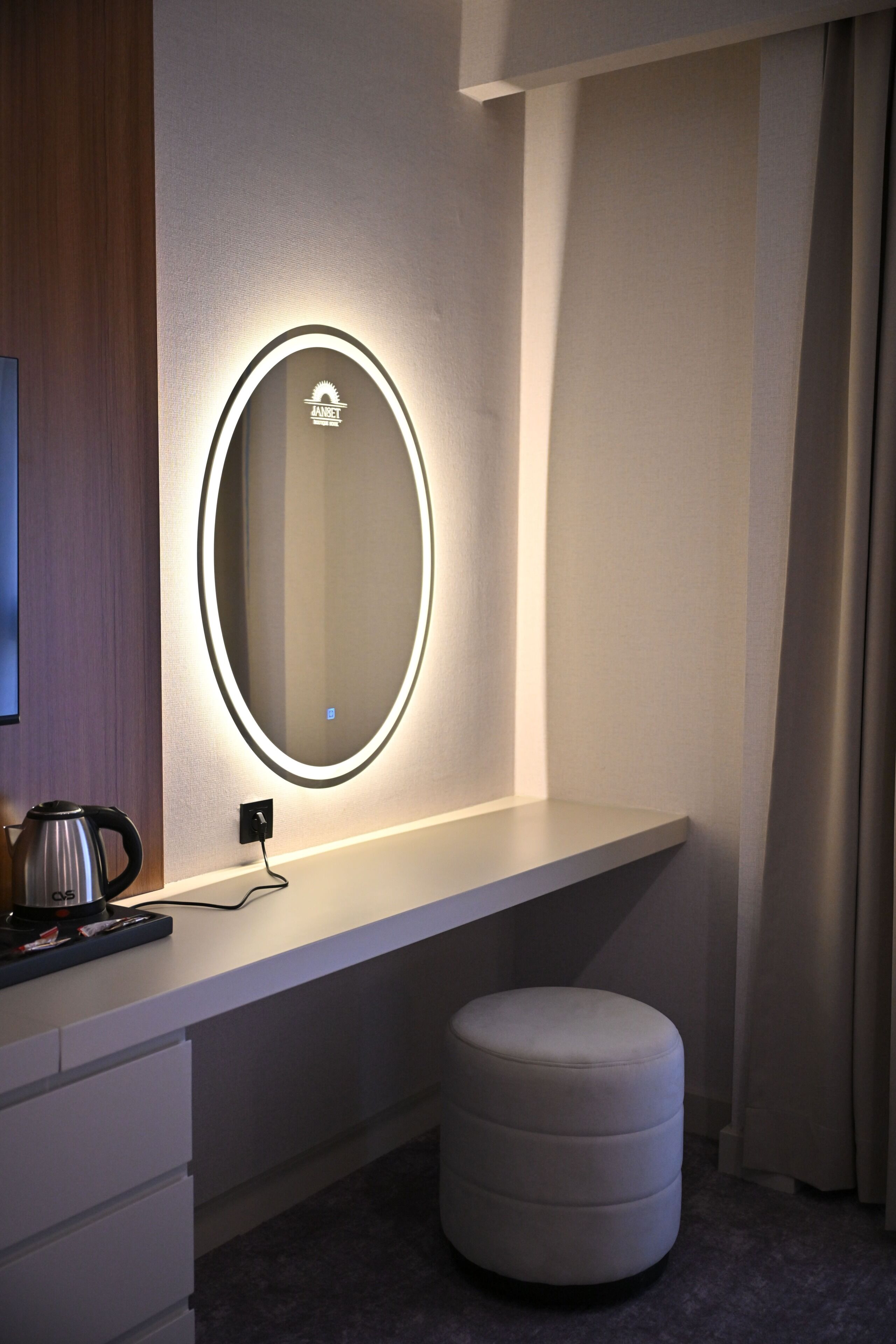 Een hotelkamer met een make-up hoek, inclusief een ronde spiegel met verlichting, een klein krukje en een flatscreen televisie.