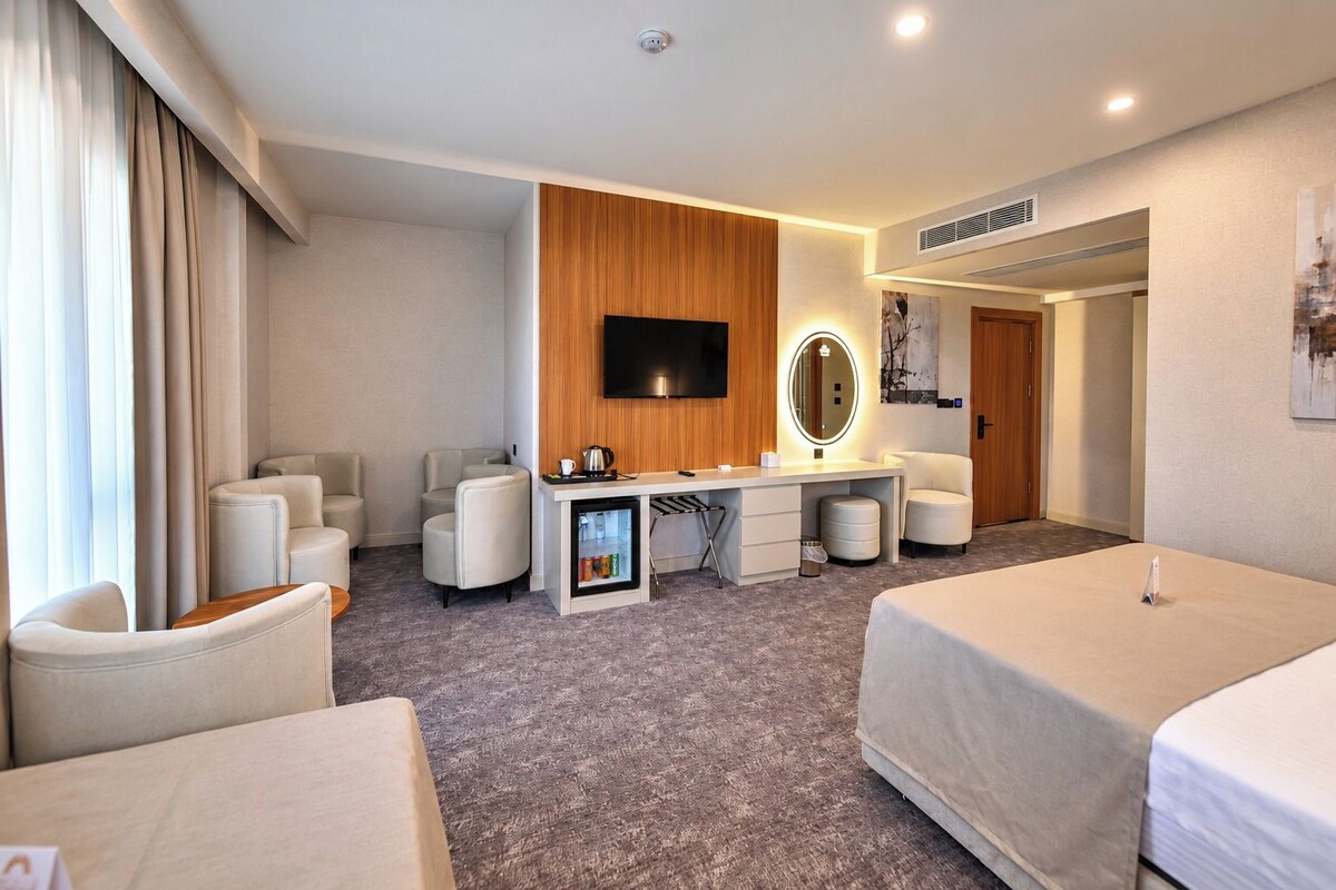 Une chambre d’hôtel comprenant un lit, un bureau avec un miroir, une télévision et un coin salon avec des fauteuils.