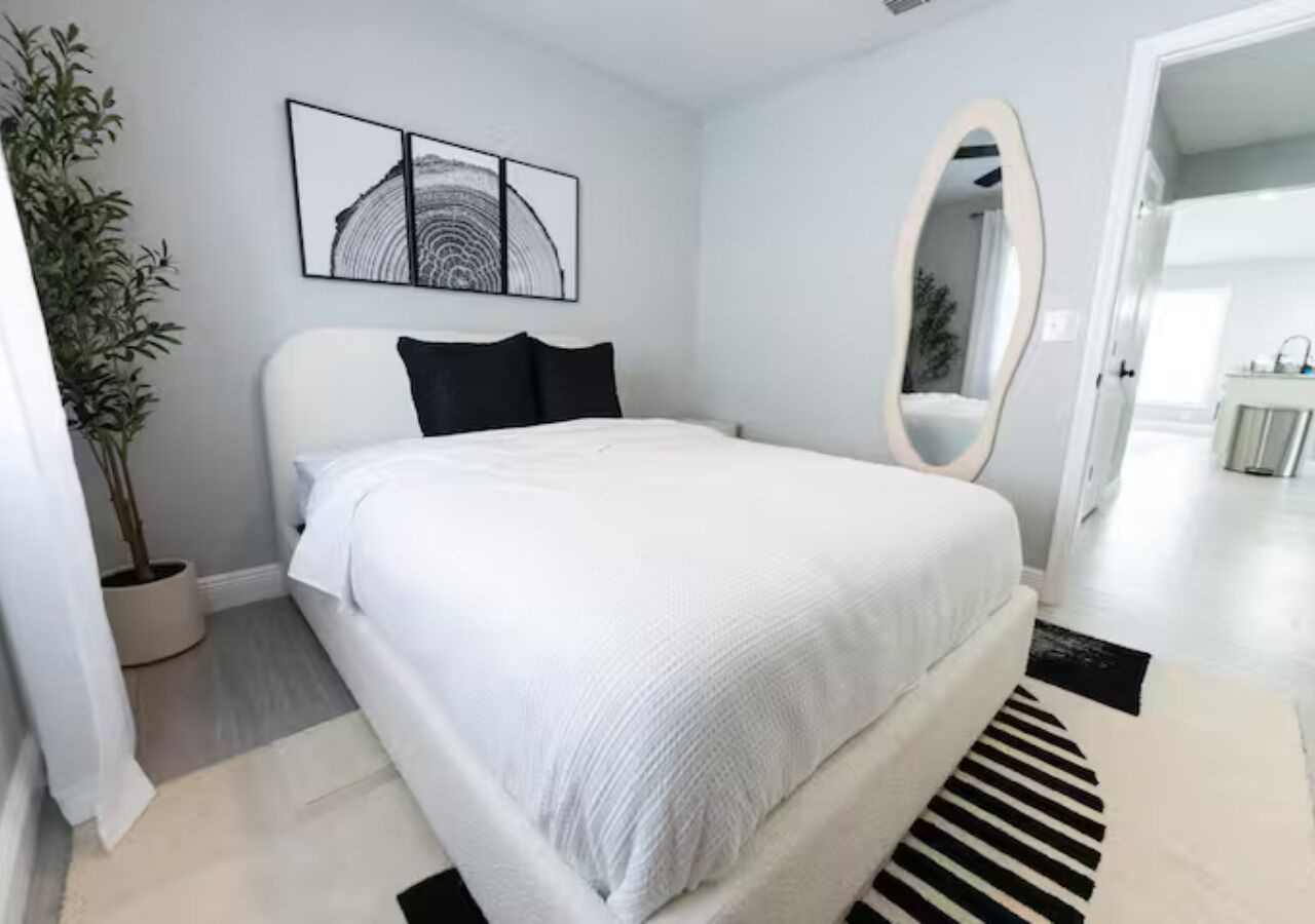 Un dormitorio moderno con cama, planta en maceta, espejo montado en la pared y una obra de arte enmarco.