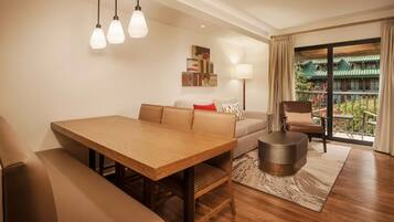 A modern living room with a wooden dining table, a sofa, a chair, and a small round table. There is a balcony with a view of greenery.