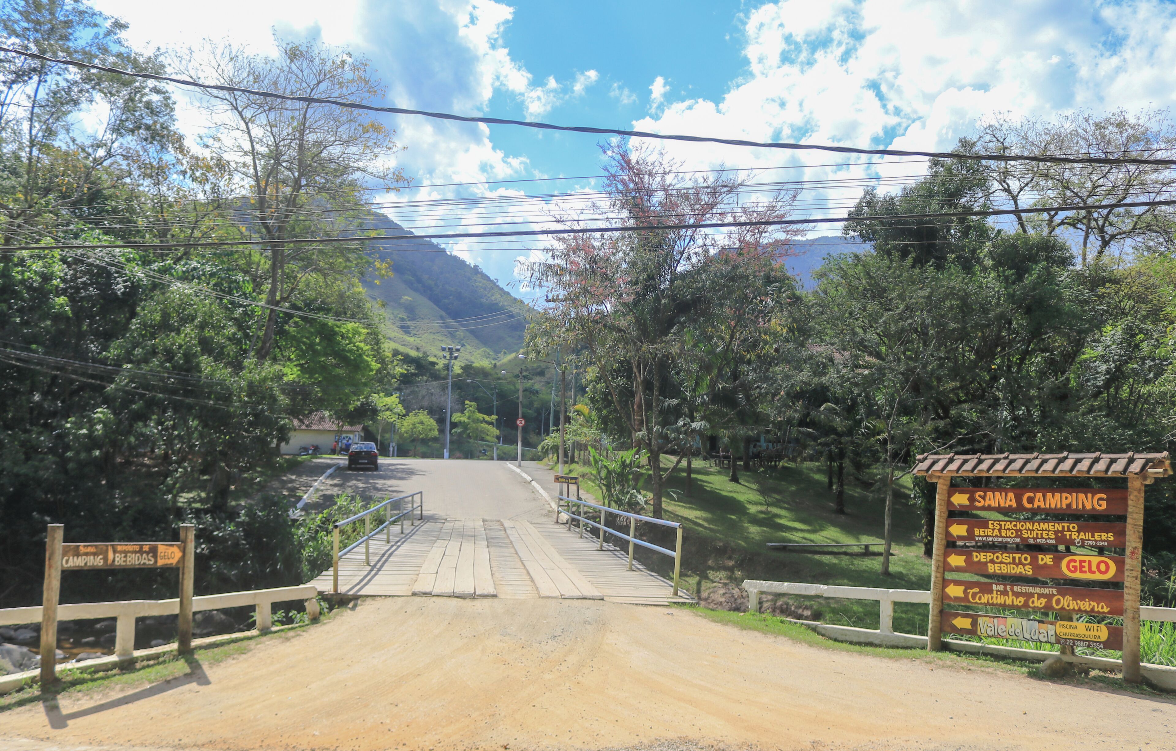 Un camino de tierra que conduce a un área de camping con un puente de madera y señales que indican servicios como un restaurante e internet WiFi.