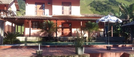 Un bâtiment à deux étages, doté d’un toit en tuiles, d’une piscine et d’un jardin agrémenté de plantes en pot.