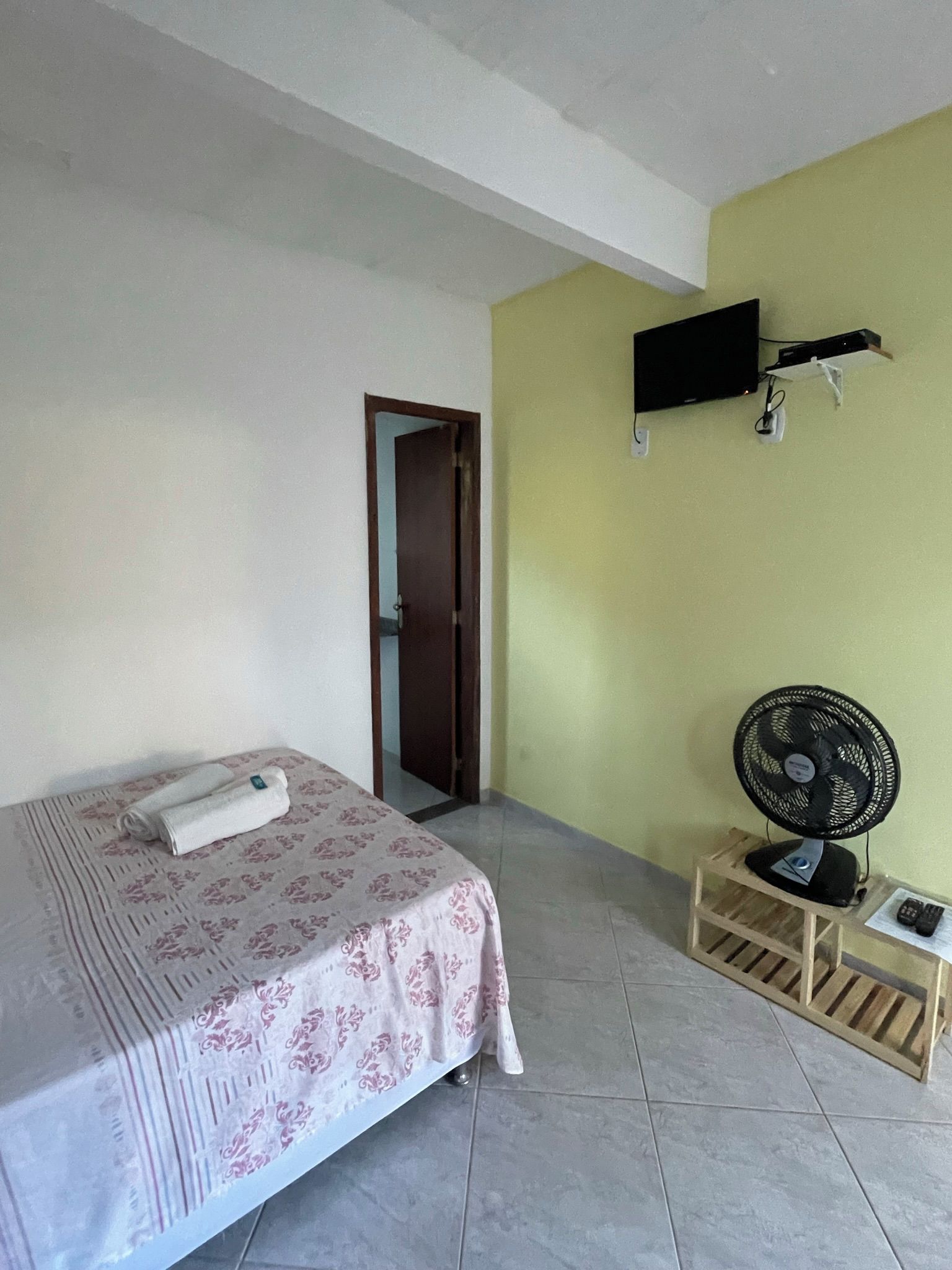 A bedroom with a bed, a wall-mounted TV, a standing fan, and a door leading to another room.
