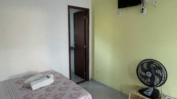 A bedroom with a bed, a wall-mounted TV, a standing fan, and a door leading to another room.