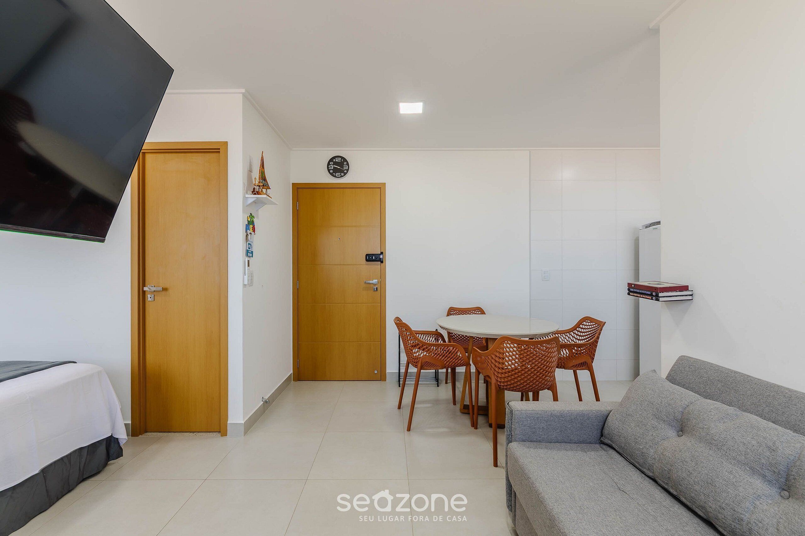 Una habitación con cama, sofá, mesa de comedor con sillas, televisor montado en la pared y un reloj en la puerta.