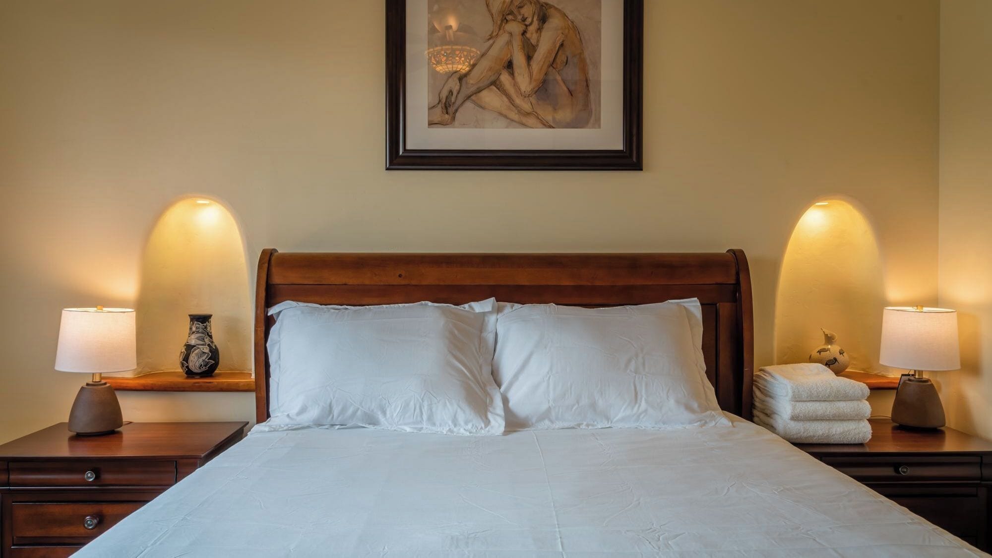A neatly made bed with white linens, flanked by two bedside tables with lamps, a vase, and a decorative item.