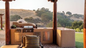 Una cocina exterior con una parrilla, un lavamanos y un horno empotrado, rodeada de pilares de madera y vistas a un paisaje montañoso.