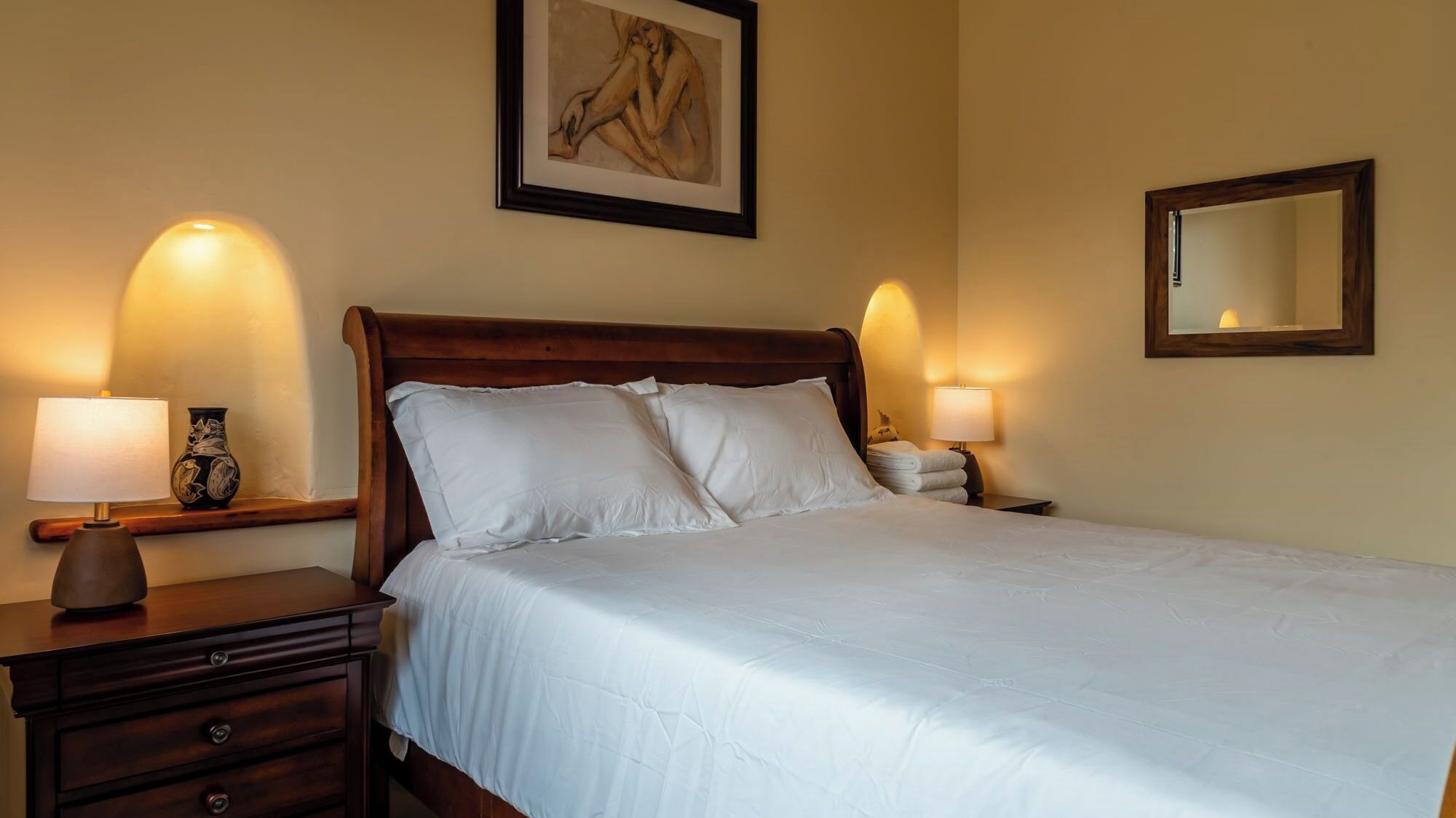 A neatly made bed with white linens, flanked by two bedside tables with lamps, a vase, and a folded towel.