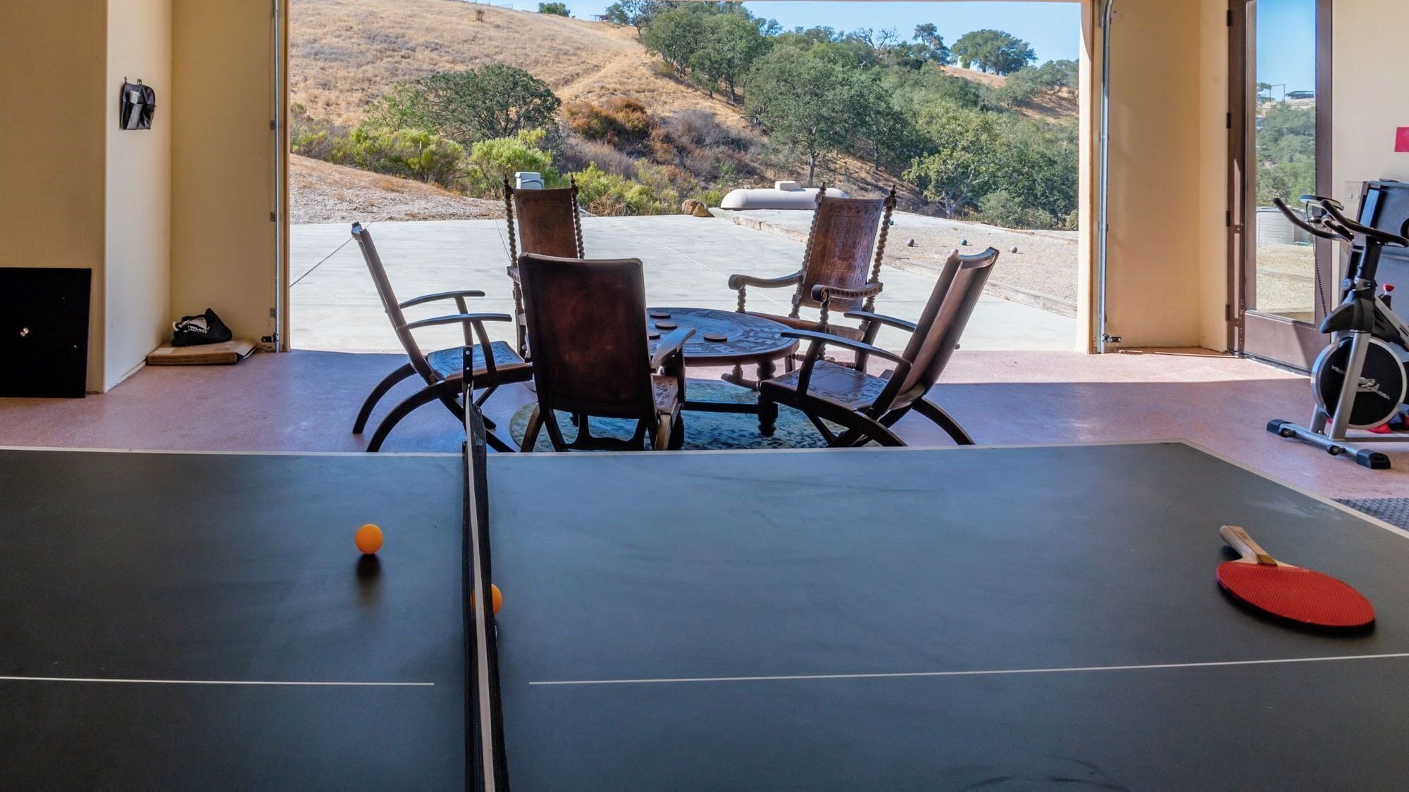 A ping pong table with chairs and a table tennis paddle, a treadmill, and a view of a hillside.
