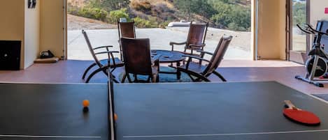 A ping pong table with chairs and a table tennis paddle, a treadmill, and a view of a hillside.