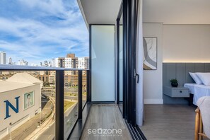 Ein Balkon mit Blick auf die Stadt, ein Bett, ein Nachttisch und ein an der Wand hängendes Kunstwerk.