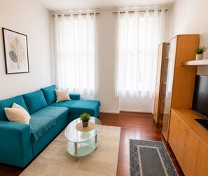 A living room with a teal sofa, a glass coffee table, a TV on a wooden cabinet, and a framed plant on the wall. - Vienna  4 Guests Double Bed + Sofa Bed Kitchen Washer WiFi (Wahring)