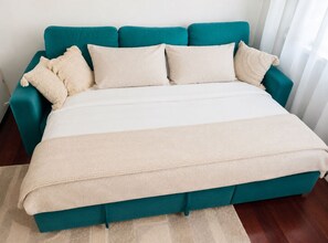 A teal sofa bed with a beige blanket and pillows, positioned against a white wall with a window. - Vienna  4 Guests Double Bed + Sofa Bed Kitchen Washer WiFi (Wahring)