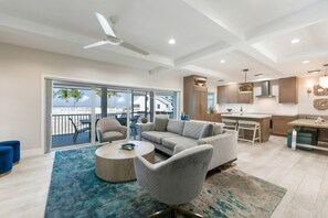 A spacious living area with a sofa, armchairs, and a coffee table. A balcony with a view of palm trees and buildings is visible through large glass doors. - Elegant Gulf-View Home | Resort Access Included (Captiva)