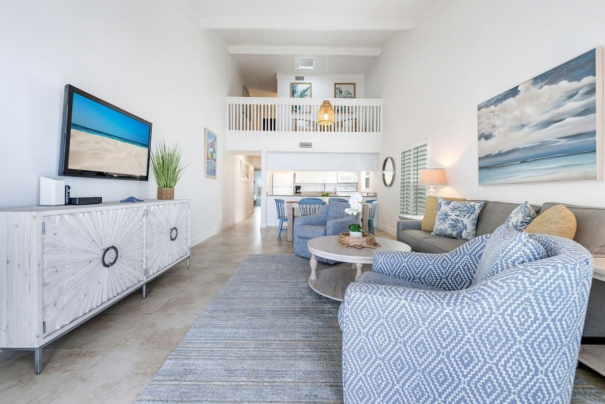 A spacious living area with a flat-screen TV, a white console, a patterned sofa, and a dining area in the background.