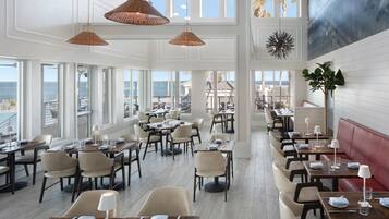 A spacious dining area with multiple tables and chairs, large windows offering a view of the sea, and hanging pendant lights.