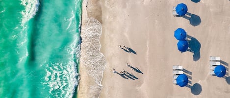 An aerial view of a beach with clear turquoise water, a sandy shore, and several rows of blue umbrellas and white lounge chairs.