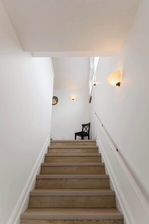 A staircase with wooden steps and white walls, leading to a landing with a chair and wall-mounted lights.