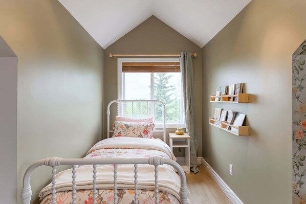 A bedroom with a sloped ceiling, a bed with a floral bedspread, a bedside table with a lamp, and a wall-mounted shelf with picture frames.