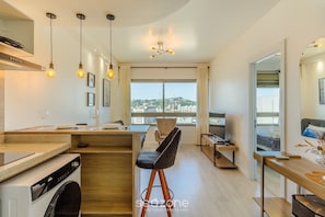 A modern kitchen with a washing machine, a dining area with a bar stool, and a large window offering a city view.