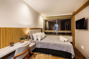A modern hotel room with a bed, a desk, a chair, a TV, and a window with blinds.