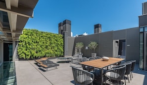 A rooftop patio with a wooden table and chairs, a green wall, and modern buildings in the background.