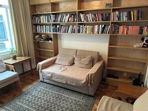 Ein gemütliches Zimmer mit einer Couch, einem Bücherregal voller Bücher, einem kleinen Tisch mit einer Pflanze und einem gemusterten Teppich.