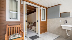 Parede exterior de madeira com um banco, uma porta que dá para um quarto com cama e mesa, e um banheiro com pia e espelho.