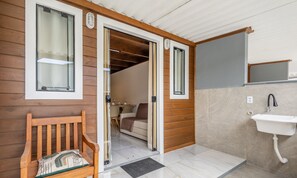 A wooden exterior wall with a bench, a door leading to a room with a bed and a table, and a bathroom with a sink and mirror. - Chalet w/ Balcony in Condo w/ Barbecue Vil0003 (Penha)