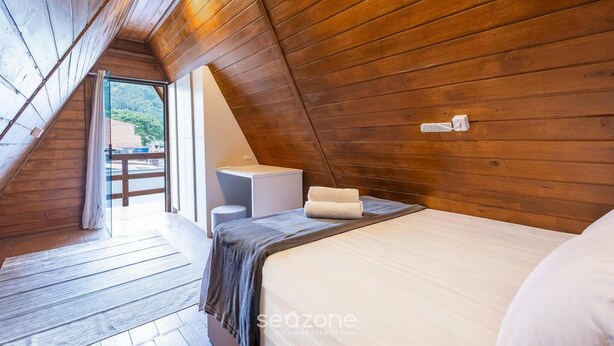 A room with wooden walls, a bed, a bedside table, a lamp, and a view of the outdoors.