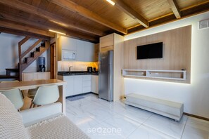 A modern kitchen with a built-in refrigerator, wooden ceiling beams, a dining area with a bench, and a TV mounted on the wall.