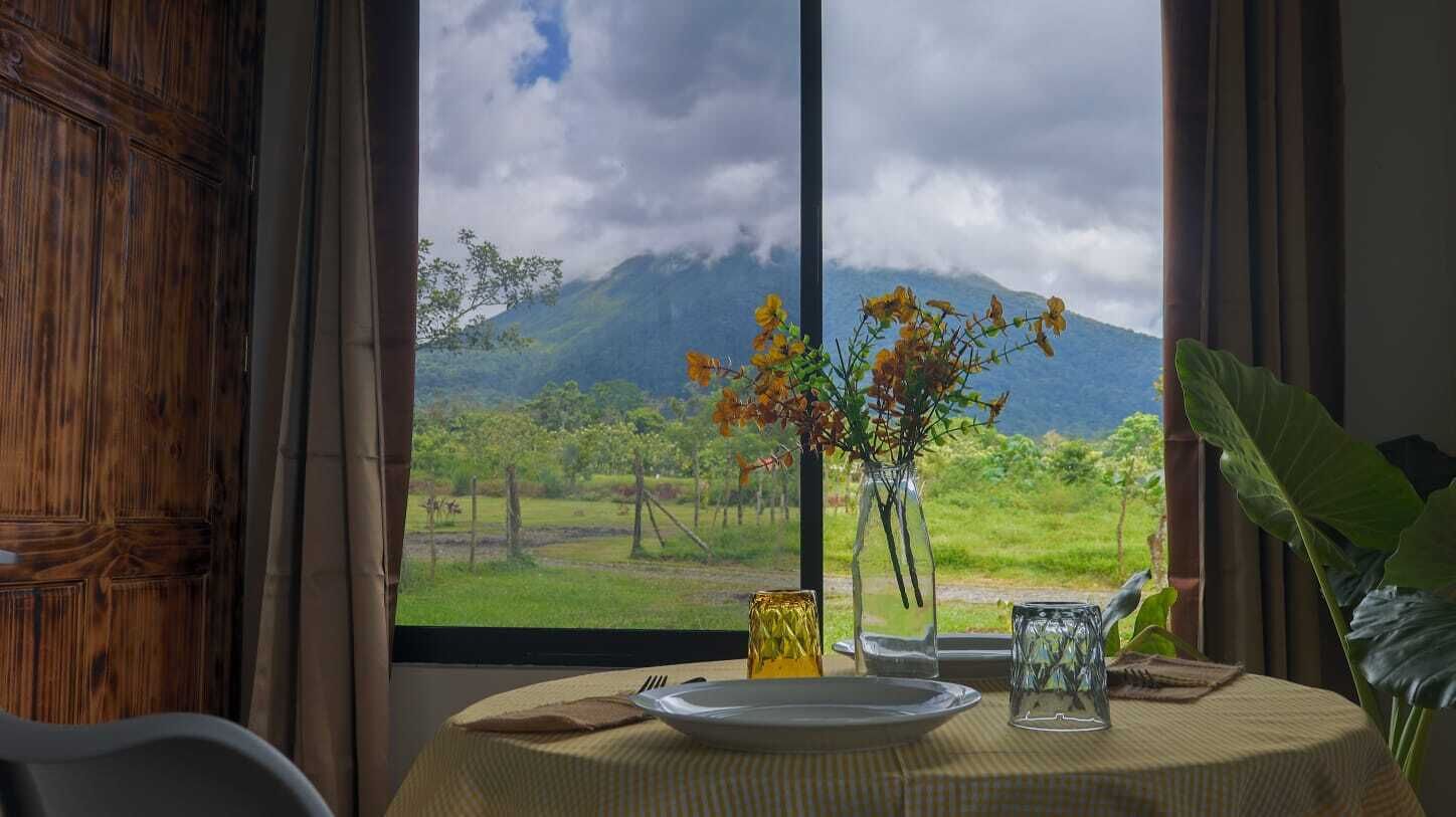 A dining table with a plate, a glass, and a vase with flowers, set against a window with a view of a mountain and greenery.