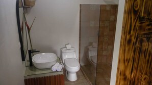 Uma casa de banho com uma pia, sanita e bidé brancos. Há um armário de madeira e uma prateleira na parede com uma cesta.